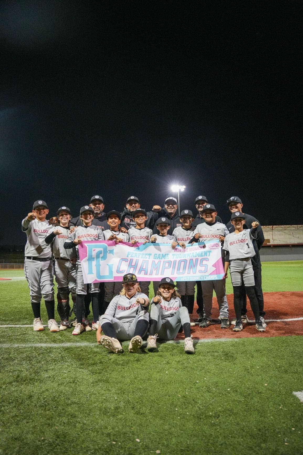 A Bulldogs youth team celebrating with a Perfect Game tournament champions banner under the lights.